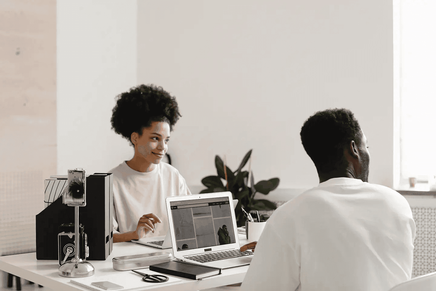 Two colleagues are collaborating at a desk, with a laptop displaying a website. The woman on the left is smiling, while the man on the right is focused on the screen. The minimalist office setting and soft lighting create a calm and collaborative mood.