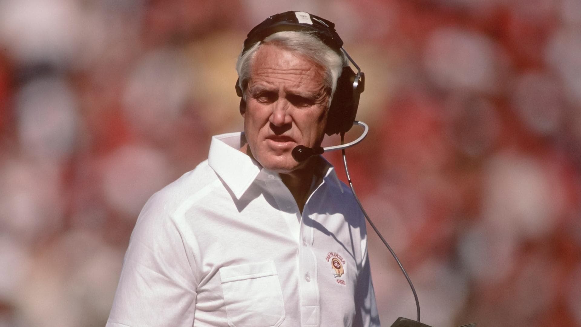 This image features a close-up of a man wearing a headset, likely a football coach. He is wearing a white collared shirt with the 'Seminole' logo, suggesting he is affiliated with Florida State University. The man has a serious expression, and the background is blurred, creating a sense of focus on the subject.
