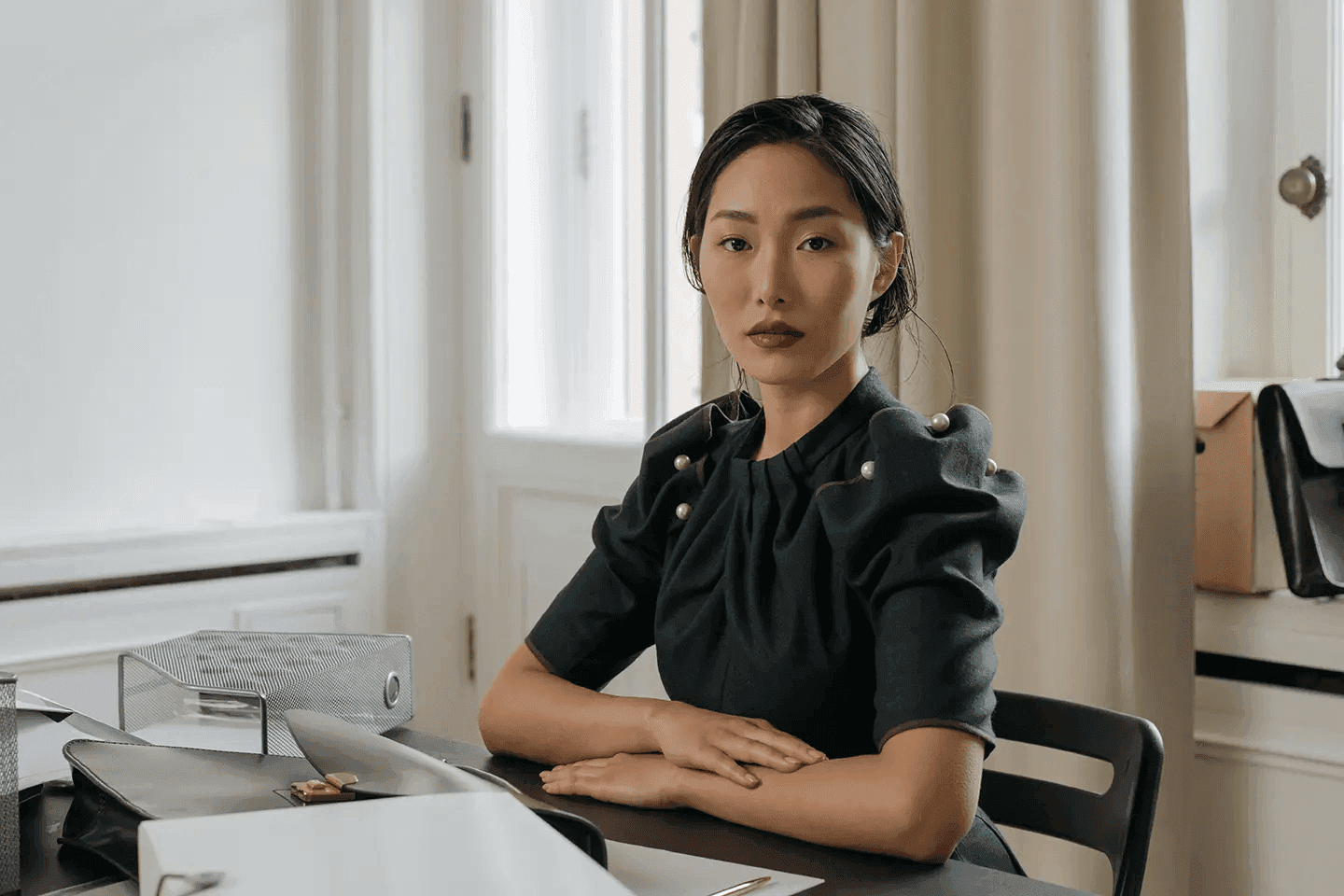 A poised woman in a stylish black dress sits at a desk, exuding confidence and professionalism. The soft lighting and neutral tones create a sophisticated and calm atmosphere. The pearls on her dress add a touch of elegance to the scene.