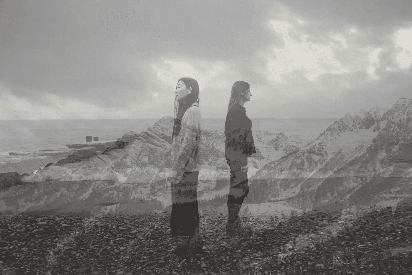This black and white image features two women standing back-to-back, overlaid with a landscape of mountains and water. The mood is contemplative and serene, with the double exposure creating a sense of connection to nature. The grayscale palette and the women's introspective poses evoke a feeling of solitude and reflection.